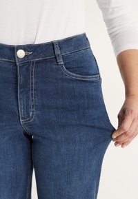 En person som bär blå denimjeans och en vit långärmad skjorta, drar i tyget på låret för att visa hur stretchy jeansen är.