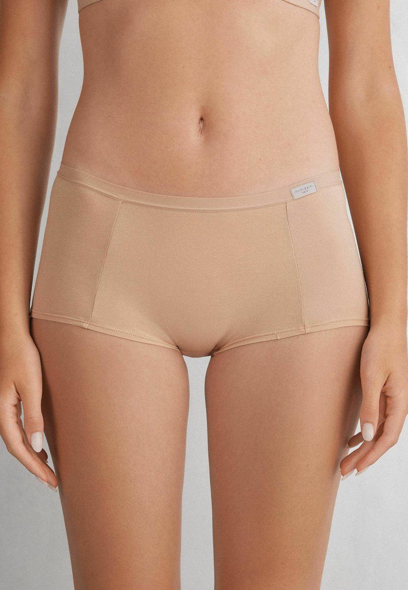 Beige hipstertrosor för kvinnor tillverkade av ett mjukt tyg, med en sömlös design och en lätt glans, samt en liten logotypetikett på midjebandet.