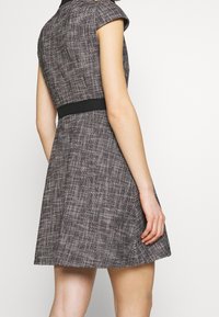 Dopasowana sukienka z rękawami w kształcie kaptura wykonana z fakturowanej tkaniny z czarną talią, z brązowym i białym wzorem tkanym. Zaprojektowana z rozszerzanym dołem.