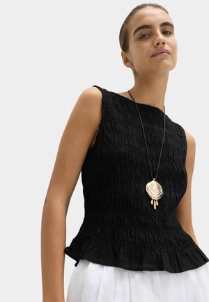 Femme portant un haut sans manches noir texturé avec un ourlet à volants et un grand collier pendentif en forme de coquillage doré sur un fond clair.