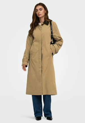Femme portant un long manteau beige avec un col noir, un jean bleu, des chaussures noires et portant un sac à épaule noir sur un fond uni.
