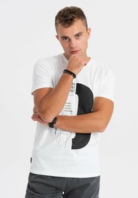 Valkoinen puuvillapaita, jossa on suuri musta graafinen kuvio, tekstitiedot ja pyöreä kaula-aukko. Paitaa täydentää mustasta helmistä tehty rannekoru.