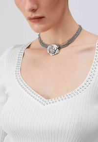 Collier pendentif fleur argentée avec plusieurs rangs de fines chaînes métalliques et accents de perles. Porté avec un haut blanc côtelé orné de clous argentés.