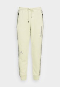 Hellgelbe Jogginghosen mit Kordelzugbund, Seitentaschen, grauen Seitenpaneelen und einem Logo in der Nähe des Knöchels. Textdetail am Bein.