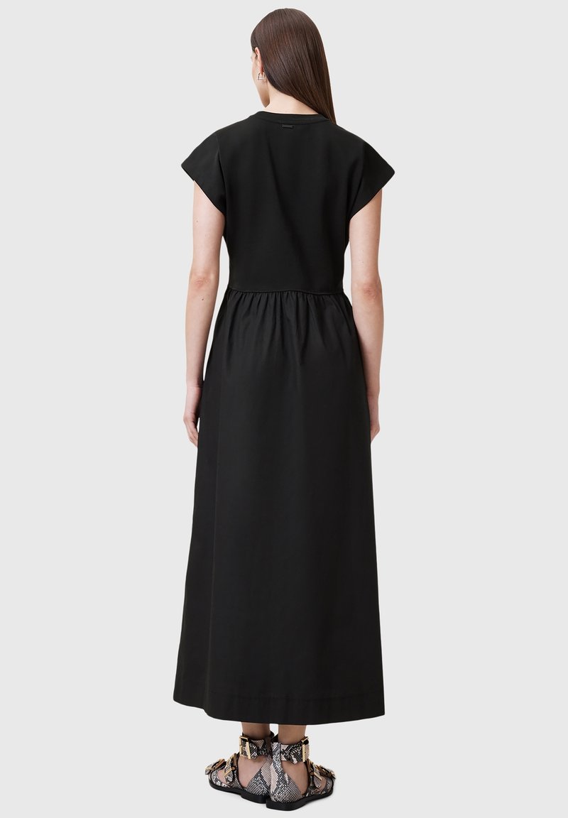Femme aux longs cheveux bruns portant une robe midi noire à manches courtes et des sandales plates à motif peau de serpent, debout devant un fond gris uni.