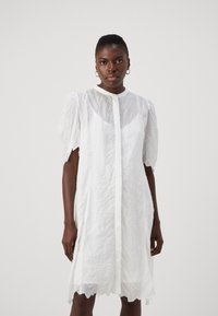 Rochie albă, semi-transparentă, cu țesătură stratificată și texturată. Are mâneci scurte, bufi și un sistem de închidere cu nasturi pe față; tivul are o margine scallopată.