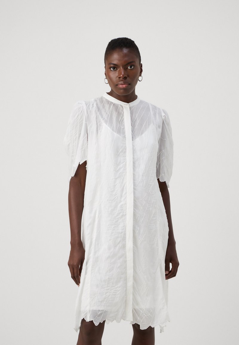 Rochie albă, semi-transparentă, cu țesătură stratificată și texturată. Are mâneci scurte, bufi și un sistem de închidere cu nasturi pe față; tivul are o margine scallopată.