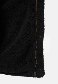 Zwarte fleecejack met een gestructureerd oppervlak, gouden ritsdetails en een marineblauwe corduroy zoom onderaan. Geen andere patronen of accenten.