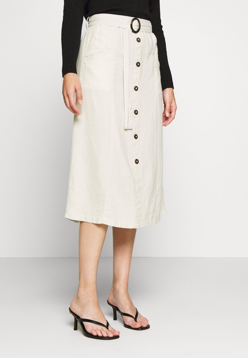 Femme portant une jupe midi blanche à boutons sur le devant avec une ceinture assortie et des sandales noires à talons, debout devant un fond uni.