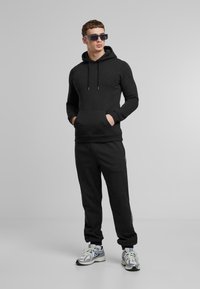 Felpa nera con cappuccio e tasca frontale, abbinata a pantaloni jogger neri con strisce bianche sui lati. Il modello indossa sneakers grigie e occhiali da sole.