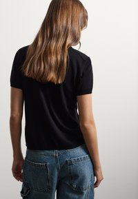 Frau mit langen braunen Haaren, die ein schwarzes Kurzarmhemd und blaue Jeans trägt, mit dem Rücken zu einer schlichten weißen Wand.