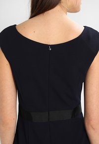 Mörkblå klänning bakifrån med rund nacke, en dold dragkedja och en satängbälte detaljer i midjan. Slät tygtextur.