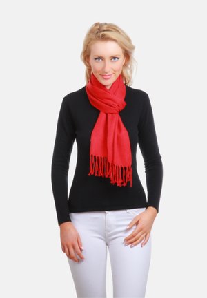 Écharpe rouge aux bords frangés, en tissu doux, drapée autour du cou. Le modèle porte un haut noir à manches longues et un pantalon blanc.