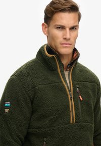 Grüner Fleece-Pullover mit einem Reißverschlusskragen, der eine strukturierte Oberfläche, kontrastierende, tannfarbene Akzente und ein kariertes Futter am Kragen aufweist.