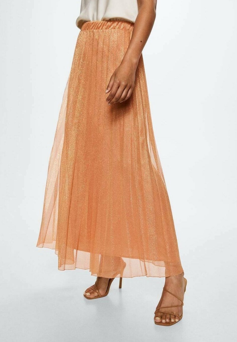Femme portant une longue jupe plissée pêche transparente en tissu scintillant et des sandales à talons hauts beiges à lanières.