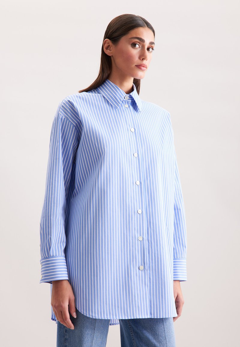 Chemise oversize rayée bleue et blanche avec un col à boutons et des manches longues. Comprend un ourlet droit et des boutons blancs sur le devant.