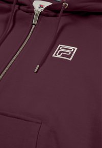 Sudadera con cremallera en burdeos, hecha de una tela suave, con un logo bordado en plata y un bolsillo frontal con textura suave y cremallera de metal.