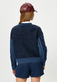 Granatowy sweter z dzianiny z teksturowanym wzorem diamentowym na rękawach, ściągacz u dołu i kontrastowe niebieskie akcenty. Noszony z ciemnymi szortami i czapką.