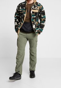 Homme portant une veste polaire camouflage, une chemise marron, un pantalon cargo vert olive et des chaussures de randonnée noires, debout les mains dans les poches.