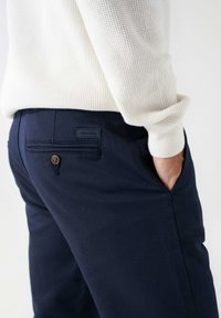 Námořnické modré kalhoty s hladkým povrchem, které mají zadní kapsu se zapínáním na knoflík a značkovou nášivku. Tkanina vypadá odolně a lehce.