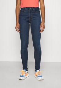 Jeans justos feitos de denim azul-escuro, com bolsos frontais, um fecho tradicional por botão, emparelhados com ténis brancos e azuis com atacadores laranja.