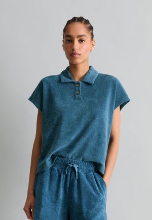 Femme aux cheveux tressés portant un polo en éponge bleu à manches courtes et un pantalon assorti avec cordon, se tenant devant un fond gris clair.