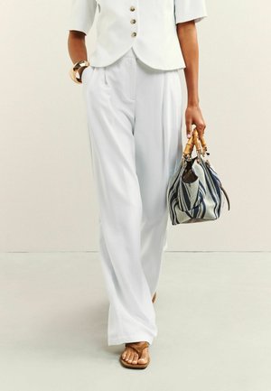 Frau trägt weiße, weit geschnittene Hose, weißes, kurzärmeliges, geknöpftes Oberteil, braune Sandalen, goldenes Armband und hält eine gestreifte Handtasche mit Bambusgriffen.