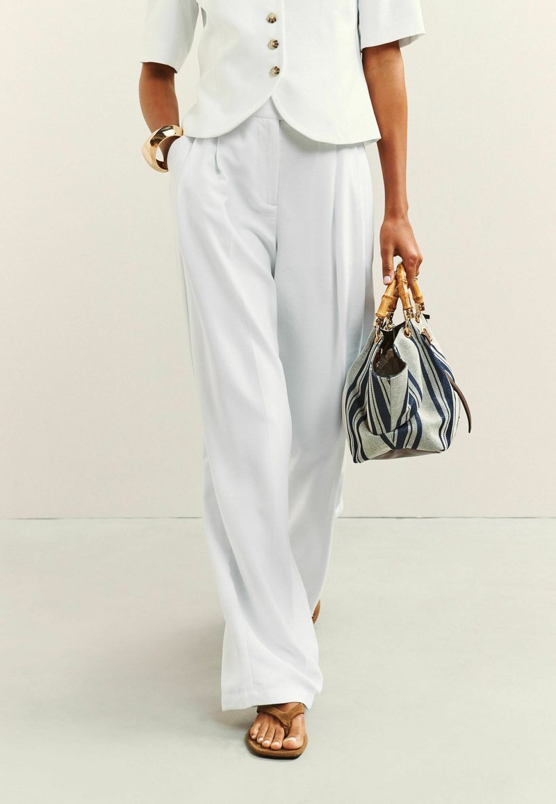 Vrouw draagt witte wijde broek, wit kortärmelig knopenblouse, bruine sandalen, gouden armband, en draagt een gestreepte handtas met bamboe handvatten.