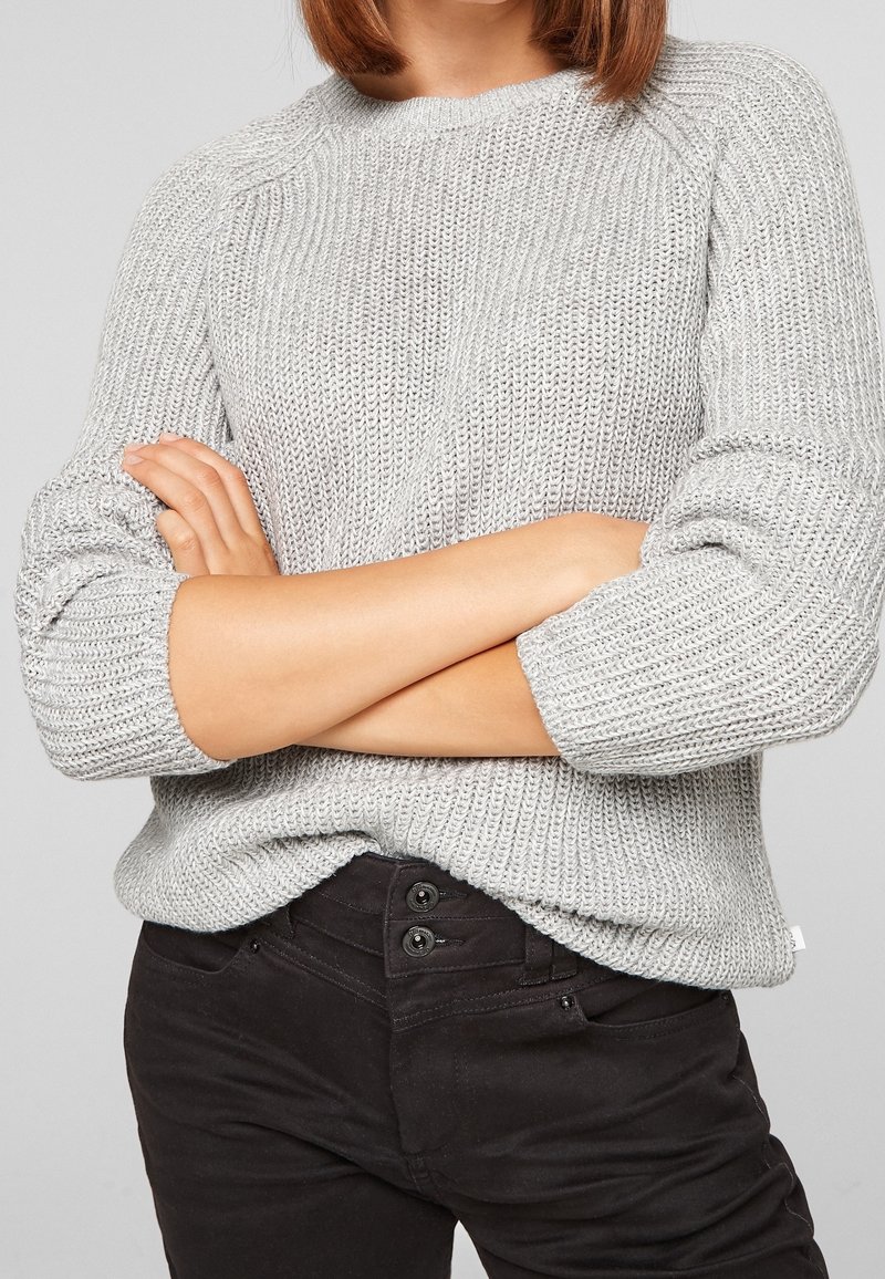 Lichtgrijze gebreide trui met een ronde hals en driekwart mouwen, gecombineerd met donkere broek met dubbele knoopsluiting.