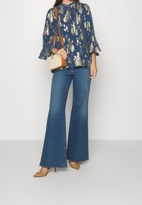 Blusa de estampado navy con detalles dorados, pantalones de mezclilla acampanados y bolso de hombro marrón, combinados con zapatos de tacón beige.