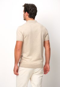 Beige kortärmad pikétröja med ribbad krage och fåll, slät textur, bärs med ljusa byxor, sedd bakifrån.