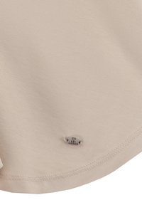 Tela beige con una superficie suave y ligeramente texturizada. Una pequeña etiqueta de logo plateada cosida en el dobladillo, que presenta "LÁRGO" en un diseño de corazón.