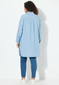 Camicia di jeans blu chiaro con bottoni, maniche lunghe, orlo curvo e pieghe sul retro, abbinata a pantaloni blu a pois e sneakers bianche.