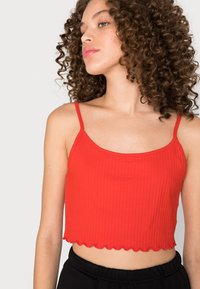 Top cropped riscado vermelho com bainha ondulada e alças finas, feito de um material suave. Estilizado com calças pretas.