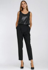 Kobieta stojąca z rękami w kieszeniach, ubrana w czarny, bezrękawnik wyszywany cekinami, czarne, dopasowane spodnie oraz czarne szpilki.