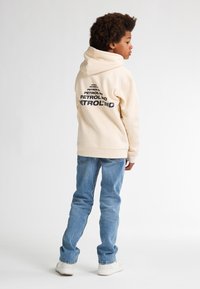 Sudadera crema con un gran logo negro en la parte posterior, combinada con jeans azules claros y zapatillas blancas. Tela suave y texturada, corte relajado.