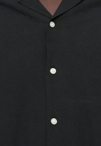 Μαύρο πουκάμισο με κουμπιά, διαθέτει χαλαρό γιακά, τρία λευκά κουμπιά στο επάνω μέρος και απαλή υφή υφάσματος.