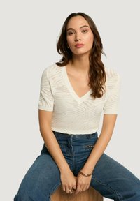 Top corto a maniche corte in maglia bianca con scollo a V e motivo testurizzato, abbinato a jeans blu a vita alta, seduto su uno sgabello di legno.