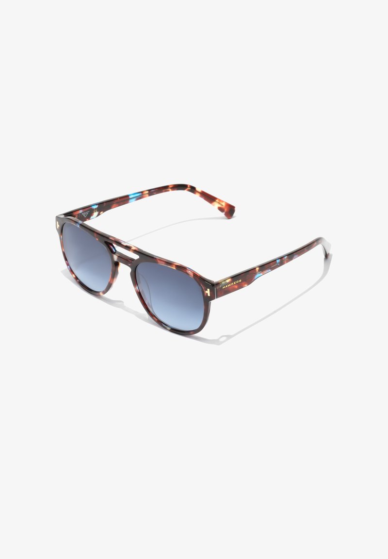 Tortie napszemüveg, fokozatos kék lencsékkel, téglalap alakú, és világosbarna díszítéssel a kereten. Arany logó részlet az oldalon.