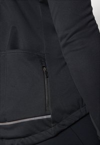 Crna atletska jakna s uskim krojem, glatkom teksturom, džepom na zip, te reflektirajućim detaljima na rubu za bolju vidljivost.