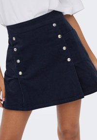 Mini-jupe en velours côtelé bleu marine avec des boutons-pression argentés sur les côtés, portée avec un haut blanc, montrant une longueur mi-cuisse et des poches avant.