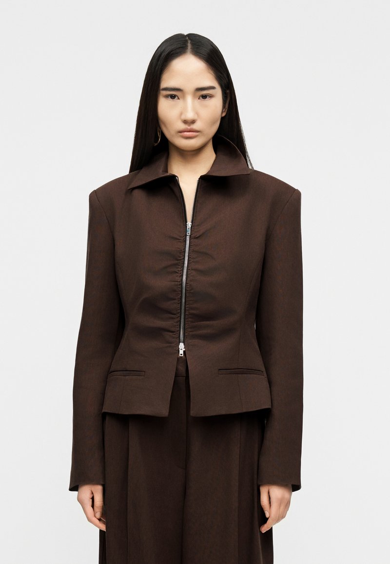 Νεαρή γυναίκα με μακριά μαύρα μαλλιά, φορώντας κ αφετί σκούρο εφαρμοστό σακάκι με εμπρόσθιο φερμουάρ και φαρδύ γιακά, στέκεται μπροστά σε απλό φόντο.