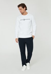 Sudadera blanca con el logotipo "SAIL RACING", combinada con pantalones oscuros de color navy y zapatillas blancas, con un corte relajado y cuello redondo.