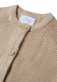 Beige gestrickte Cardigan mit Rundhalsausschnitt, zwei Knöpfen und gerippten Bündchen. Das Etikett trägt die Aufschrift "Stellon & Friends", Größe 62/68. Sichtbare Struktur des Garns.