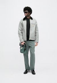 Siva jakna na patent s crnim ovratnikom, crna majica, zelene hlače i mala zelena torbica; nosi sunčane naočale i crne tenisice.