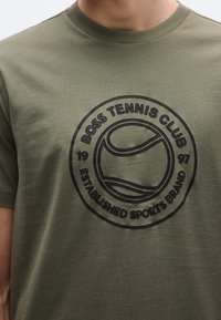 Olívazöld pamut póló fekete hímzett kör alakú logóval, rajta "BOSS TENNIS CLUB" felirattal és egy teniszlabda grafikával. Rövid ujjú.