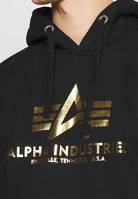 Felpa nera con cappuccio decorata con un logo in oro metallico e accenti a strisce. Coulisse con occhielli in argento; tessuto morbido e strutturato.