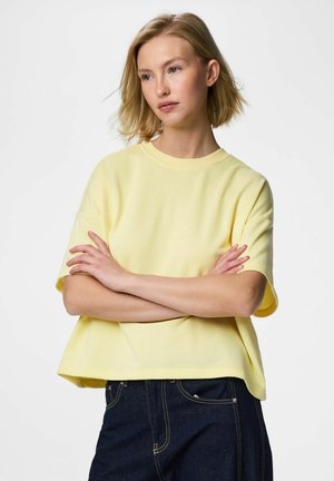 Giovane donna con capelli biondi corti, indossa una camicia gialla a maniche corte e larga e jeans scuri, con le braccia incrociate, che guarda di lato.