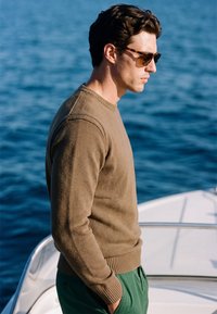 Homme portant des lunettes de soleil, un pull marron et un pantalon vert, debout sur un bateau, regardant vers l'eau, les mains dans les poches.
