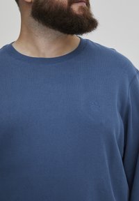 Sudadera azul de tejido suave, con escote redondo y un pequeño detalle de logo en el pecho. Textura lisa y corte relajado.